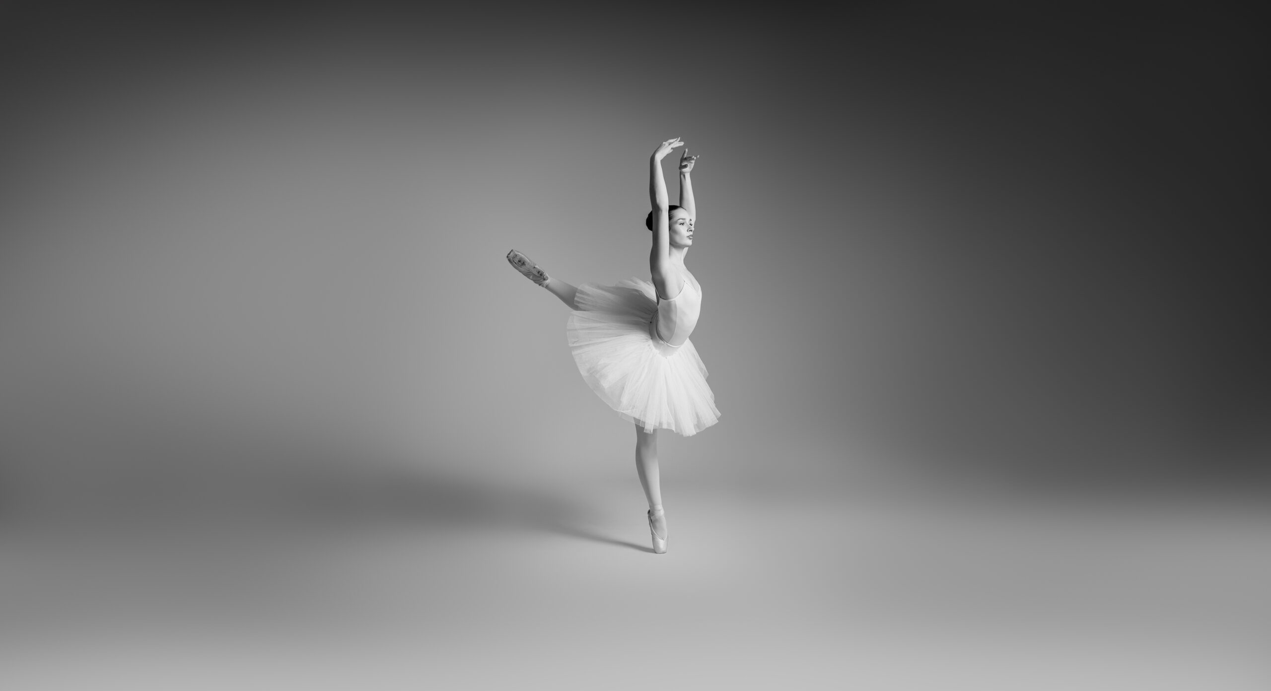 Eine Ballerina in einem weißen Tutu steht auf Spitzen, ein Bein hinter sich ausgestreckt und die Arme anmutig erhoben, und tanzt in einem sanft beleuchteten, minimalistisch gestalteten Studio – ein atemberaubendes Beispiel für die Ballettfotografie von Matzios Tanzfotografie Fineart Stuttgart.