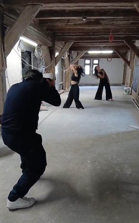 Kostas Matzios in schwarzer Kleidung fotografiert zwei Tänzerinnen, die in schwarzen Outfits unter einer Holzbalkendecke in einem noch unfertigen Innenraum des Fruchtkastens in Herrenberg posieren, und hält dabei dynamische Momente während eines Tanzfotografie-Shootings für eine örtliche Tanzschule fest.