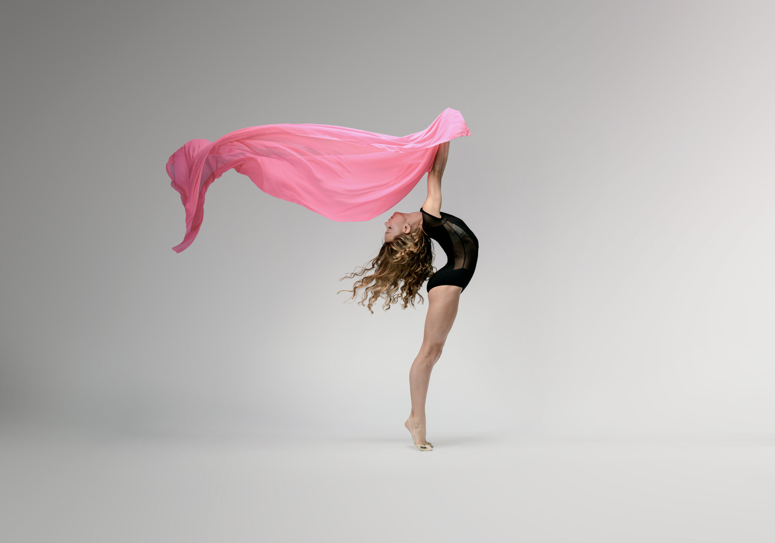 Eine Tänzerin in einem schwarzen Trikot springt anmutig in die Höhe und hält dabei ein wallendes rosa Tuch über ihren Kopf – in einer minimalistischen, hellgrauen Studiokulisse, die die Eleganz der Rhythmischen Sportgymnastik bei einem inspirierenden Fotoshooting einfängt. Ballett Fotograf Kostas Matzios