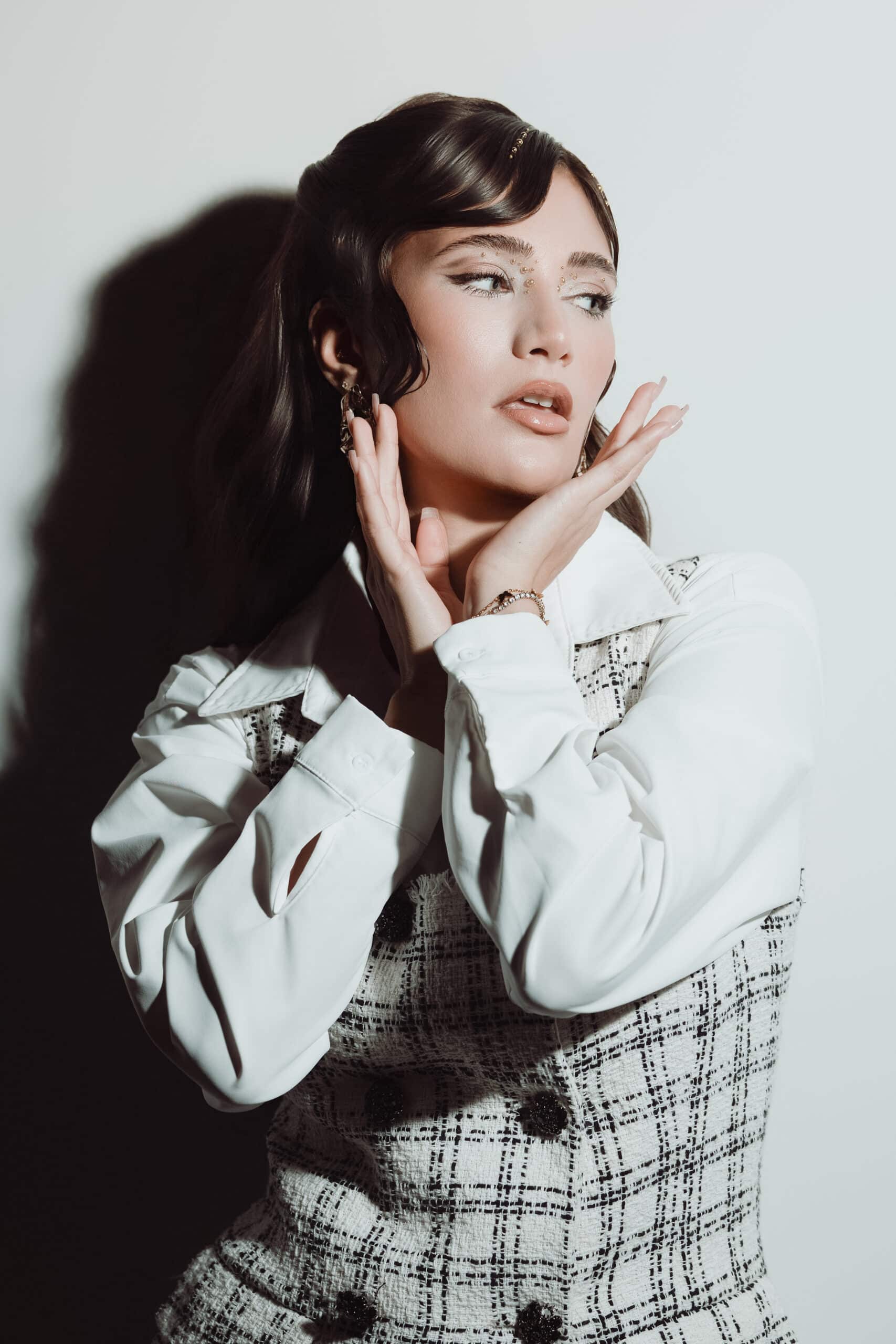 Eine Frau mit gestyltem dunklem Haar posiert vor einem weißen Hintergrund und trägt ein weißes Hemd sowie eine gemusterte Weste. Aufgenommen im unverkennbaren Fine-Art-Stil von Matzios Fotoshooting hält sie ihre Hände nahe an ihr Gesicht und blickt mit nachdenklichem Ausdruck zur Seite – ein wahrhaft eindrucksvolles Porträt und eines der besten Beispiele aus Stuttgart.