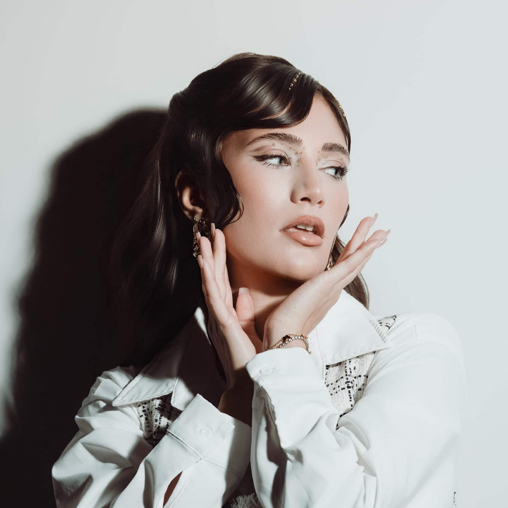Eine Frau mit gestyltem dunklem Haar posiert vor einem weißen Hintergrund und trägt ein weißes Hemd sowie eine gemusterte Weste. Aufgenommen im unverkennbaren Fine-Art-Stil von Matzios Fotoshooting hält sie ihre Hände nahe an ihr Gesicht und blickt mit nachdenklichem Ausdruck zur Seite – ein wahrhaft eindrucksvolles Porträt und eines der besten Beispiele aus Stuttgart.